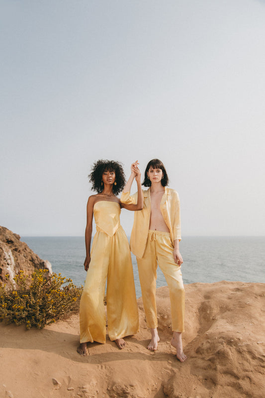 two models holding hands against beach background wearing matching silk blazer and pants in lemon yellow