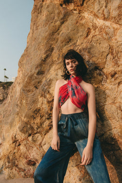 model in leaning against beach rocks wearing silk tie dye scarf as top and silk wide leg pants in sapphire blue color with unique markings
