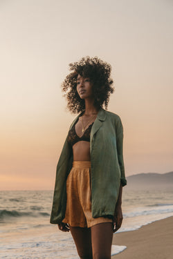 model on the beach wearing moss green silk blazer and amber brown silk shorts with a black bralette