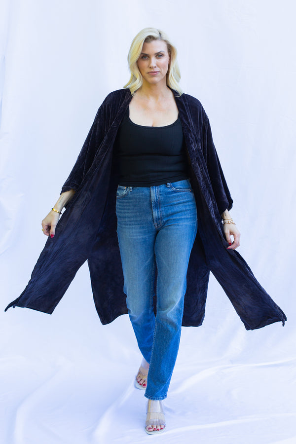 Blonde woman walking forward wearing velvet duster in midnight (deep purple) color. Duster is blowing in the breeze showing side slits. 