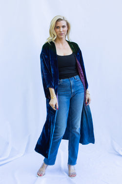 Blonde woman standing with weight shifted to one side wearing velvet duster in emerald night. She is also wearing blue jeans, a black tank, and clear heels. 