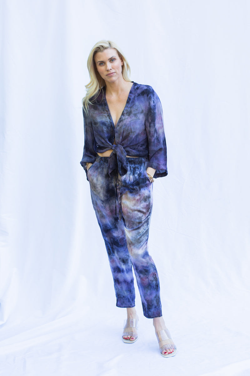 Front view of woman wearing matching set with hands in pant pocket. Featuring cosmica dharma pants in purple tie dye color.