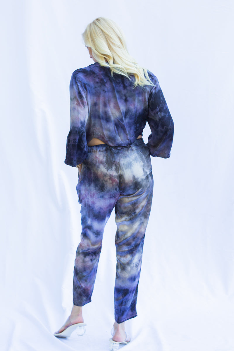 Back view of woman wearing matching set with hands in pant pocket. Featuring cosmica dharma pants in purple tie dye color.