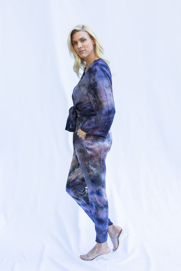 Side view of woman wearing matching set with hand in pant pocket. Featuring cosmica dharma pants in purple tie dye color.